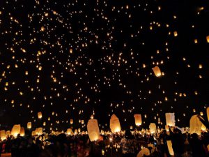 a crowd of people release paper lanterns with candles inside that make them float into the sky. the dark sky is full of small glowing lanterns floating away.